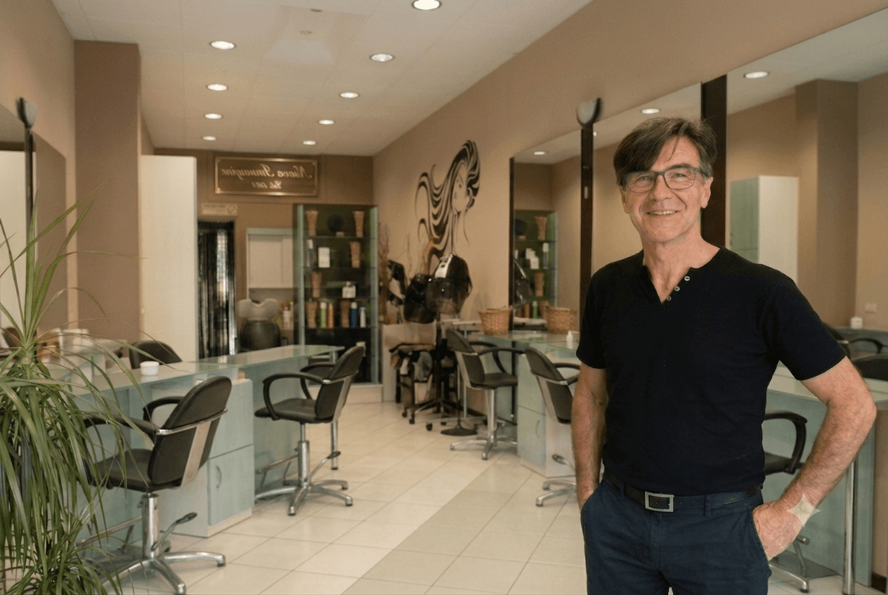 Parrucchiere sorridente con occhiali in un salone luminoso con postazioni di lavoro e specchi.